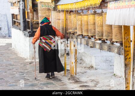 Lhasa, Tibet, China - November 15, 2019:  Old Tibetan woman walks past prayer drums and touches the drum with her right hand. Ancient traditional pray Stock Photo