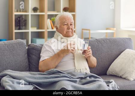 Sick senior man with a glass of water in his hand suffers from a severe cold sitting on the couch. Stock Photo