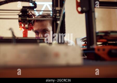 Focused young man watching modern 3D printer creating red plastic model in studio Stock Photo