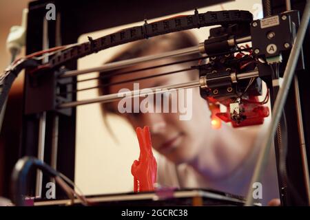Focused young man watching modern 3D printer creating red plastic model in studio Stock Photo