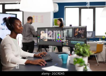African american video editor wearing headset editing movie using post production software working in start up company office. Black creator processing video footage on computer with dual displays Stock Photo