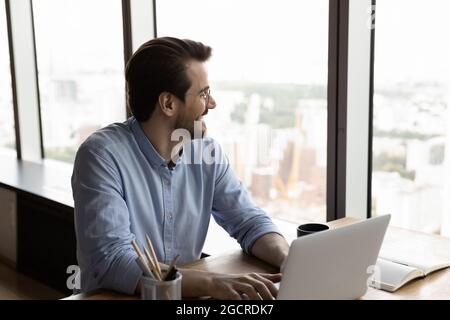 Happy confident thoughtful businessman using laptop, looking out office window Stock Photo