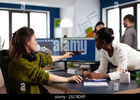Diverse women game developers creating game interface sitting in startup creative production company pointing at pc displays. Workers developing online graphics video games with modern technology Stock Photo