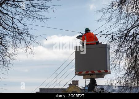 Electrician in a lift platform wearing protective work clothes changing public street lights on a sunny day. Stock Photo