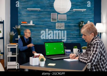 Confident senior woman using laptop Elderly woman working on computer with green screen and husband holding tv remote control. Stock Photo