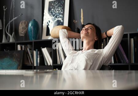 Woman with satisfied expression at desk. Arms up and folded behind her head in modern office. Stock Photo