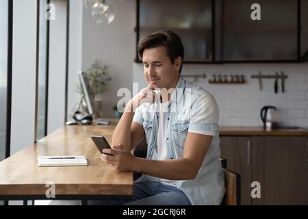 Happy thoughtful millennial guy using app, online service on smartphone Stock Photo