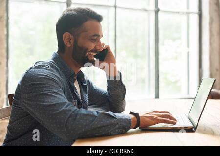 Optimistic and cheerful Indian freelancer guy talking on smartphone and using laptop sitting at desk in office, multiracial eastern man typing on keyboard during phone conversation Stock Photo