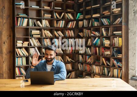 Positive smiling Indian freelancer guy using laptop for virtual meeting at office or library, holding virtual meeting, multiracial eastern male employee waving and greeting online interlocutors Stock Photo