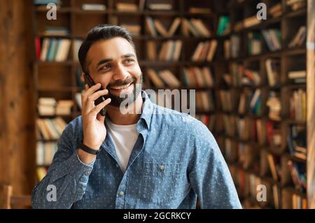 Smiling Indian businessman in smart casual shirt talking on the smartphone, eastern man has pleasant phone conversation stands in modern office with bookshelves on background, looks away and laughs Stock Photo