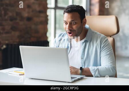 Excited surprised indian dark-haired man in casual shirt staring at laptop, shocked happy middle-eastern male entrepreneur received good deal, emails with job offer Stock Photo