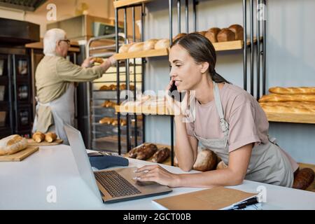 Busy female in apron using laptop and talking to clients on the phone by workplace Stock Photo