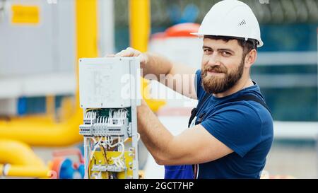 Electrician in a helmet and overalls works adjusts the equipment looks into the camera and smiles. A positive portrait of a kind Caucasian worker with a beard at work. Energy Industry Service. Advertising photo for the site. Stock Photo