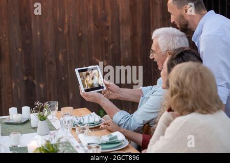 Senior man holding tablet while his family communicating with friends in video chat after dinner Stock Photo