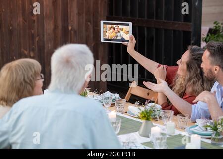 Young and senior couples communicating with their friends or relatives in video chat during dinner Stock Photo
