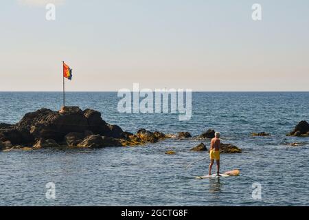 Senior man stand up paddle boarding (SUP) by the rocks on the seashore of the old fishing village Boccadasse, Genoa, Liguria, Italy Stock Photo