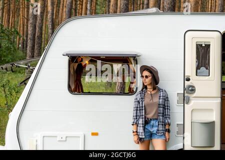 Pretty female traveler in casualwear and sunglasses standing by house on wheels Stock Photo