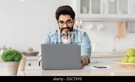 Remote business. Happy arab freelancer man working with laptop in kitchen, typing on computer, enjoying online job Stock Photo