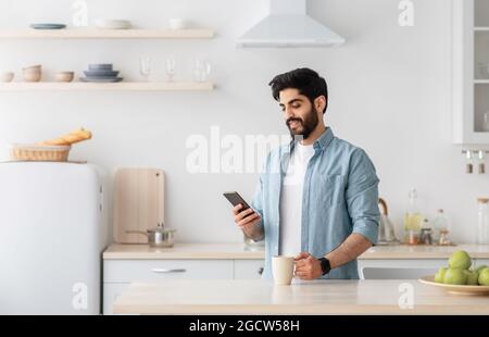 Happy young arab man using smartphone and drinking morning coffee in kitchen, empty space Stock Photo