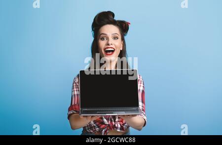 Excited pinup woman in vintage outfit showing laptop with empty screen on blue background, mockup for ad or website Stock Photo