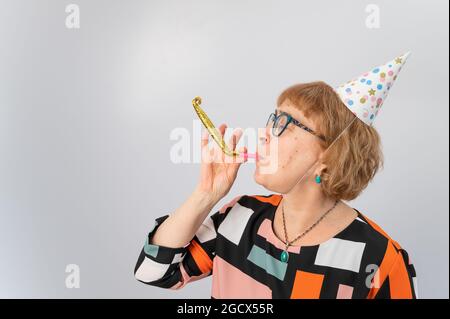 Portrait of a smiling elderly woman in a festive cap holding a whistle tongue on a white background Stock Photo