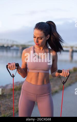 Young woman doing biceps exercise by expander at riverbank, vertical Stock Photo