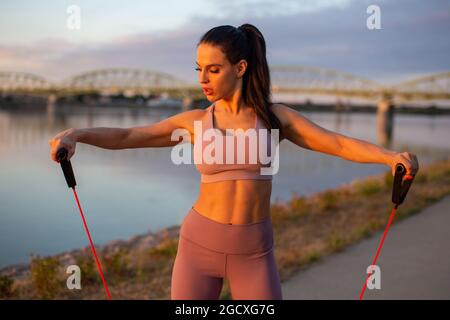 Young woman doing biceps exercise by expander at riverbank, horizontal Stock Photo
