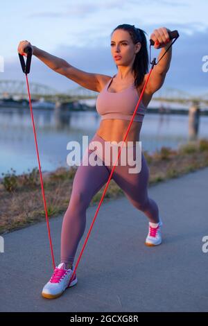 Young woman doing triceps exercise by expander at riverbank Stock Photo