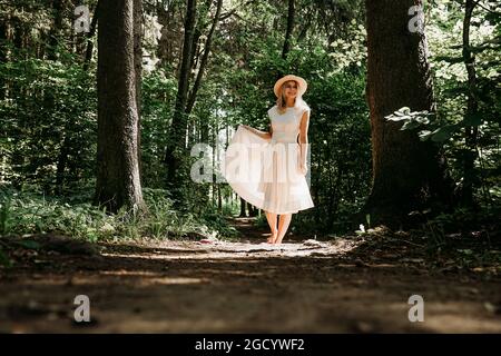 A girl in a white dress and hat walks through a summer park or forest. Fairy forest. Full-length photo of happy woman Stock Photo