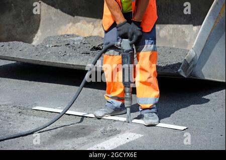 Worker preparing lay for asphalting the road using pneumatic road breaker, standing in front of a shovel of loader. Stock Photo