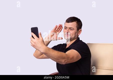 The man in black t-shirt sitting on a chair using hearing aid talking by the video call on the cellphone and waving. Stock Photo
