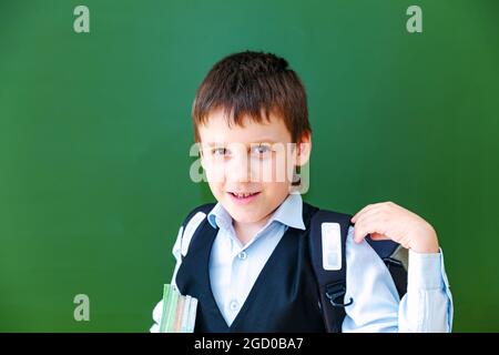 Funny schoolboy grimaces near the green school board in the classroom. Elementary school child with bag and books. Back to school. Stock Photo