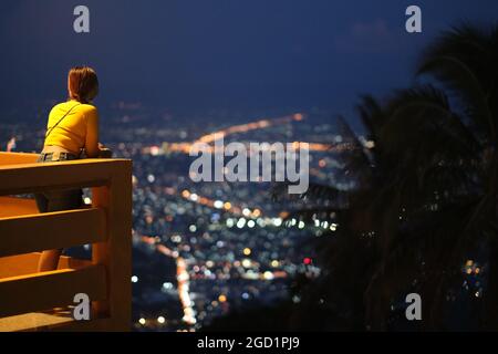 Young woman looks around night lights of a big city (Chiang Mai) while standing on a balcony high above Stock Photo