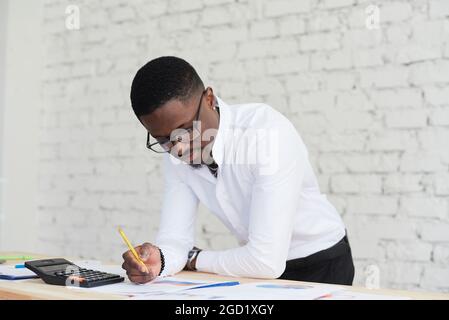Young african executive in shirt stands at his desk in the office, working with papers and graphs. Stock Photo