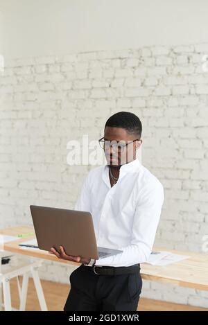 Concentrated African American man uses laptop, notepad and business papers in a modern office. Young black male freelancer stands and works in coworki Stock Photo