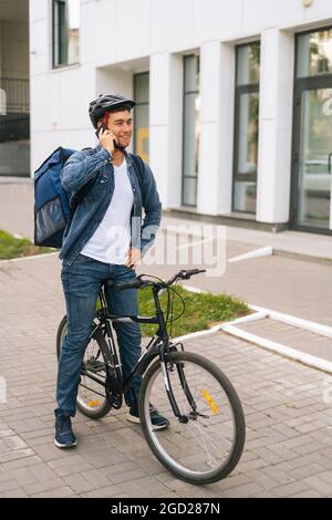 Cheerful Caucasian courier male with thermal backpack talking on mobile phone to customer, looking for address to deliver order. Stock Photo