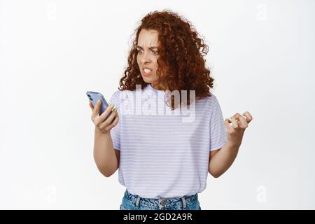 Angry and annoyed redhead woman yelling at smartphone, looking outraged at mobile phone screen, complaining, standing over white background Stock Photo
