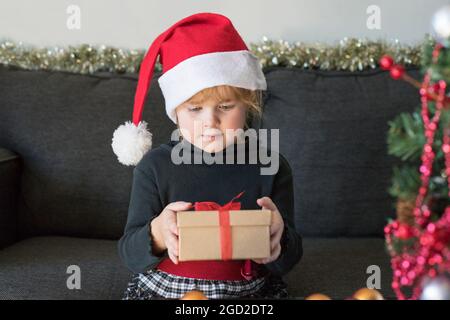 Child with christmas presents. Christmas and New Year celebration. Child holding gift box, wearing dress and santa hat, decorated artificial fir tree Stock Photo