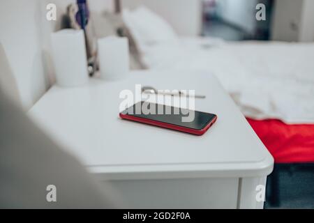 Black smartphone in a red case on the bedside table. Women watch on the table in the bedroom against the background of the bed. Close up Stock Photo