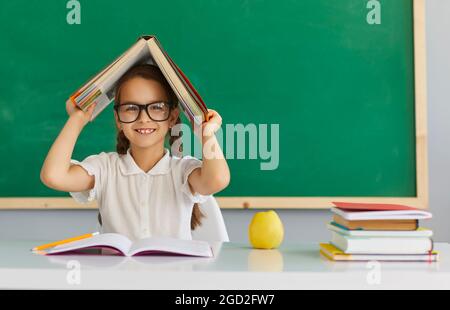 Schoolgirl in glasses holds a book over her head while sitting at a lesson against the background of the school blackboard. Stock Photo