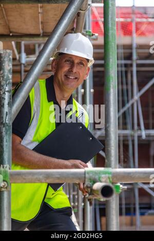Male builder foreman, smiling happy construction worker or site manager holding a clipboard, wearing a white hard hat and hi vis vest Stock Photo