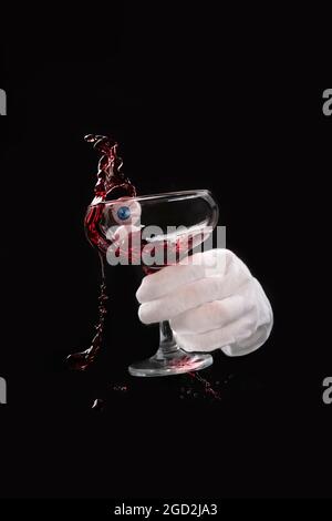 Waiter's hand in a white glove shaking a glass with red wine with eyes, splashes and waves of drink on a black background, halloween concept, scary ho Stock Photo