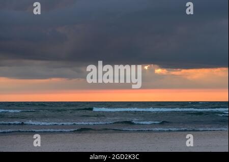 dark clouds at sunset by the sea with small waves and the sun scattered behind the clouds only leaving an orange stripe on the horizon Stock Photo