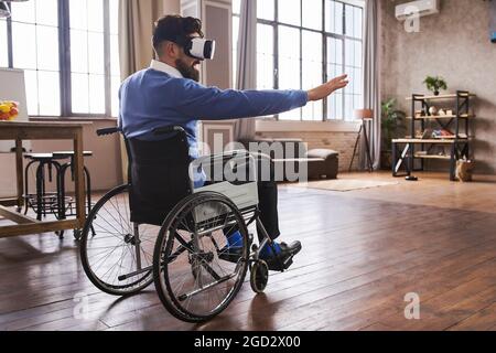 Man in a wheelchair using a VR headset and trying to touch virtual objects Stock Photo