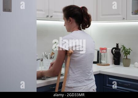 Woman on crutches washes the dishes in the kitchen. Temporary limitation of the physical capabilities of a person with a broken leg. Stock Photo