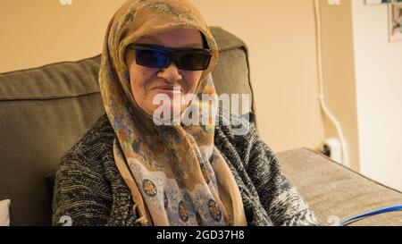 Senior Muslim woman with 3D glasses watching TV. She is happy and enjoying the show. Concept of senior woman using technology Stock Photo
