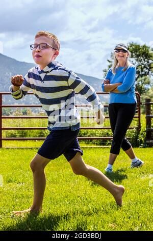 Laughing woman watching young boy playing & running in grassy yard Stock Photo