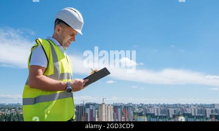 Smiling man in white helmet at construction site writes notes on clipboard Stock Photo