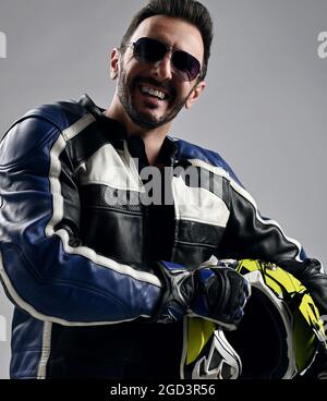 Happy smiling bearded man biker in motorcycle leather jacket and gloves stands holding his helmet and laughs Stock Photo