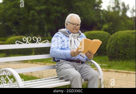 Happy relaxed senior man resting reading an interesting book sitting on a park bench. Stock Photo